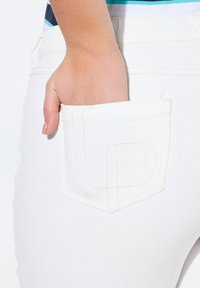 Jeans de mezclilla blanca con detalles en costura amarilla. Mano parcialmente metida en el bolsillo trasero, mostrando el diseño minimalista del bolsillo y su costura.