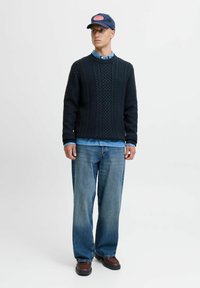 Pull en maille torsadée bleu marine, chemise en denim bleu clair, jean bleu clair coupe ample, chaussures marron, casquette bleu marine avec logo rouge. Fond minimal.