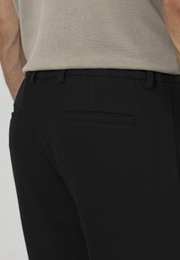 Zwarte maatpakbroeken met een gestructureerde afwerking, voorzien van een enkele achterzak en riemlussen, gestyled met een lichtgrijs bovenstuk.