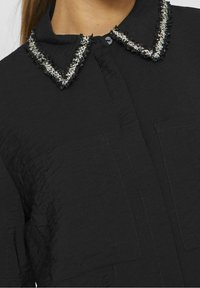 Black shirt featuring a textured fabric, a collar with white and black fringed accents, and two front pockets. Unique stitching detail visible.