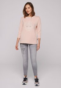 Felpa rosa chiaro con un design grafico rotondo e dettagli argento, abbinata a jeans skinny grigi con stampe testuali e sneakers chunky.