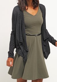 Žena v olivově zelených bezrukávových šatech s černým páskem a tmavě šedém kardiganu s volnými rukávy, který je rozepnutý vpředu.