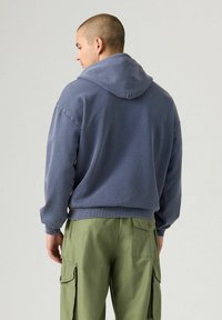 Hombre con cabello corto vestido con una sudadera azul y pantalones cargo verdes, de perfil frente a un fondo claro y sencillo.
