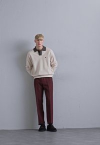 Lichtbeige fleece trui met een donkere kraag, gecombineerd met bordeaux op maat gemaakte broeken. Zwarte schoenen; minimalistisch ontwerp met een klein logo.
