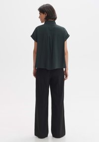 Blusa corta verde scuro con maniche corte, piega sul retro, colletto e vestibilità ampia, abbinata a pantaloni neri a gamba larga con vita alta.