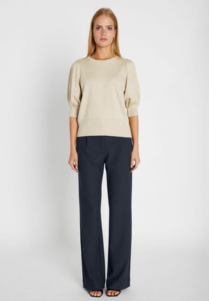 Kobieta w beżowym swetrze z bufkami na rękawach i czarnych spodniach z szerokimi nogawkami, stojąca z rozluźnionymi ramionami na jednolitym białym tle.