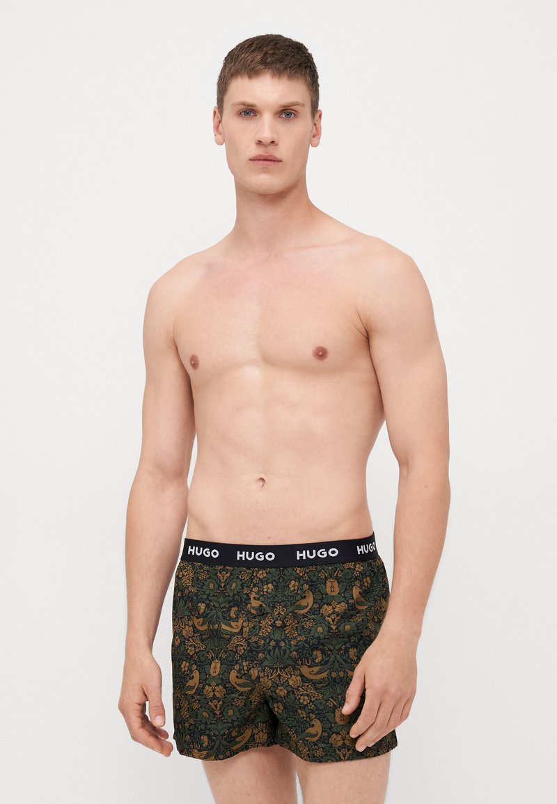 Herrenboxershorts mit einem schwarzen elastischen Bund, der mit der Marke "HUGO" versehen ist. Der Stoff ist dunkelgrün mit einem aufwändigen goldenen und braunen Blumenmuster.