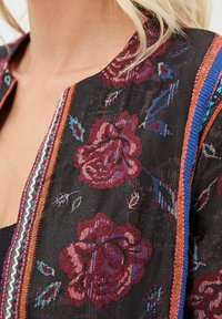 Acercamiento a una tela negra con intrincado bordado floral rojo y un colorido borde rayado en una prenda usada por una persona con cabello rubio.