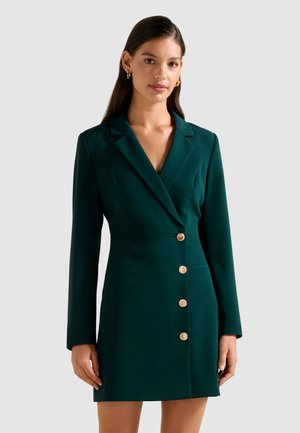 Femme portant une robe blazer taillée vert foncé avec des boutons dorés, se tenant contre un fond clair uni.