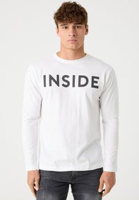 Camiseta de manga larga blanca con el texto negro en negrita "INSIDE" en el pecho. Confeccionada en algodón suave y con un cuello redondo.