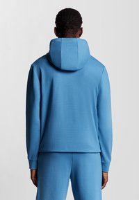 Felpa azzurra chiara con un ampio cappuccio strutturato, tessuto morbido, maniche lunghe e una vestibilità comoda, abbinata a pantaloni della tuta blu coordinati.