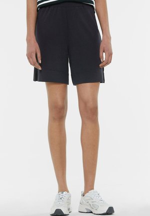 Person trägt schwarze knielange Shorts und weiße Turnschuhe und steht vor einem schlichten Hintergrund.