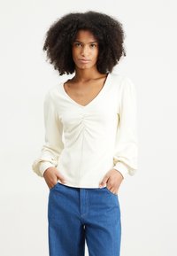 Cream-colored long-sleeve top with a V-neck and gathered front details, paired with blue denim pants. Smooth fabric and relaxed fit.