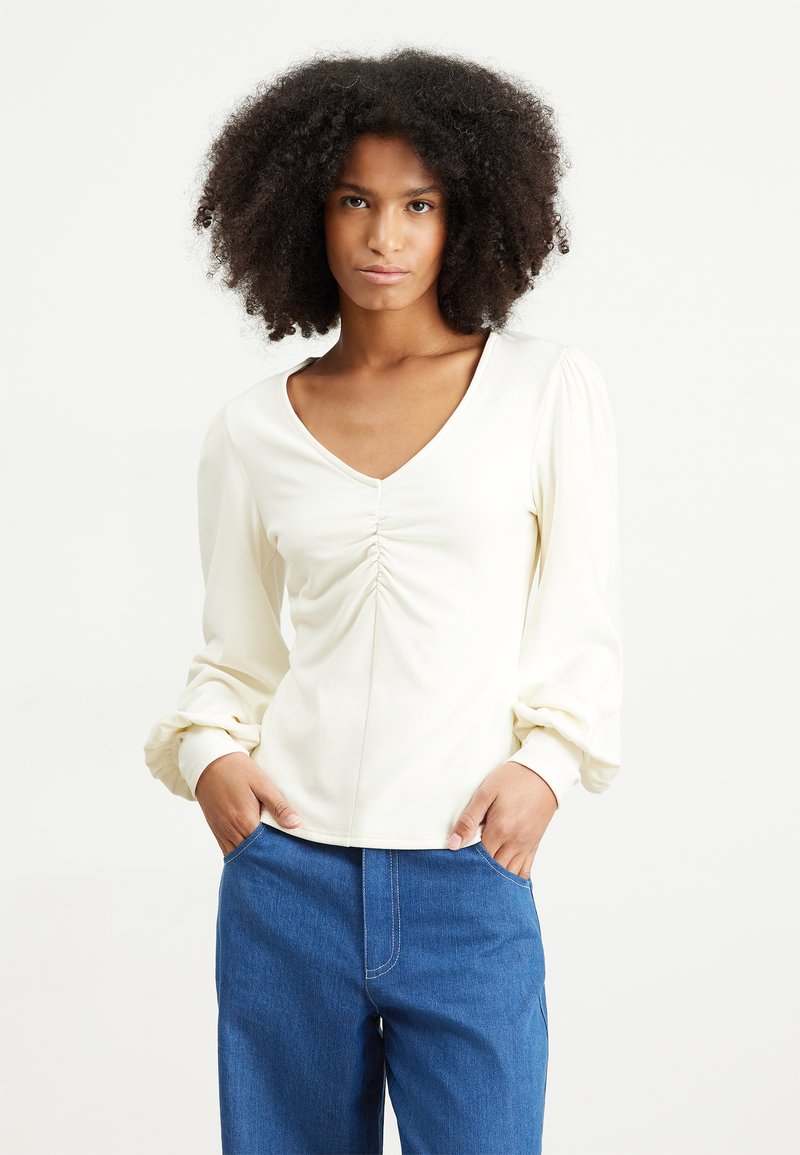 Cream-colored long-sleeve top with a V-neck and gathered front details, paired with blue denim pants. Smooth fabric and relaxed fit.