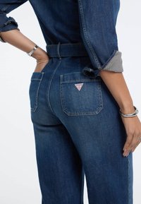 Jeans in denim di colore blu scuro, a vita alta, con gambe larghe e un logo a triangolo sulla tasca posteriore. Presentano maniche arrotolate e anelli argentati.