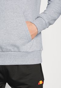 Sweat à capuche en coton gris avec une poche frontale, des poignets et un ourlet côtelés. Associé à un pantalon noir avec un logo multicolore sur la jambe.