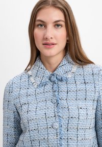 Lichtblauwe tweedjas met een textuurpatroon in een raster, rafelige kraag en decoratieve knopen. Gladde afwerking met subtiele geweven accenten.