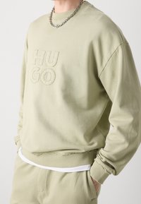 Beige sweatshirt met het doorgestikte "HUGO" logo, bijpassende broek, zichtbaar wit ondershirt, zilveren ketting, handen van persoon in de zakken.