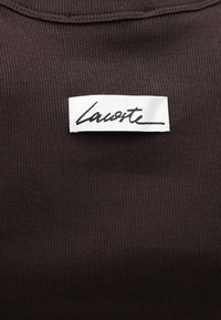 Fond en tissu côtelé marron avec une étiquette rectangulaire blanche comportant le texte en écriture cursive noire "Lacoste" cousu au centre.
