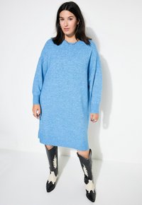 Ylkeä sininen neulemekko, jossa on pitkät hihat, ribbed hihansuut ja sivuhaalareita, yhdistettyinä musta-valkoisiin cowboy-kengiin, joissa on lehtikuvio.