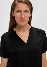 Zwarte blouse met een v-halsontwerp, korte mouwen en subtiele plooien op de schouders. Gladde textuur en minimalistische stijl.