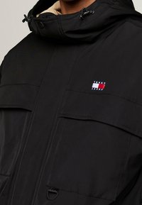 Czarna kurtka z kapturem, posiadająca duże przednie kieszenie. Materiał wydaje się gładki o matowym wykończeniu. Na piersi widoczne logo Tommy Jeans.