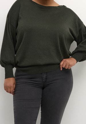 Kvinde iført mørkegrøn langærmet sweater med puffede ærmer og tætsiddende manchetter, kombineret med mørkegrå smalle jeans, hånd på hoften.