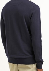 Un homme portant un pull en maille de couleur bleu marine uni à manches longues et un pantalon beige clair, vu de dos, se tenant devant un fond blanc.