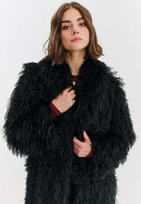 Černý kabát z umělé kožešiny s hrubou, chlupatou texturou, s širokým límcem a dlouhými rukávy, nošený přes burgundské tričko. Vysoký límec.