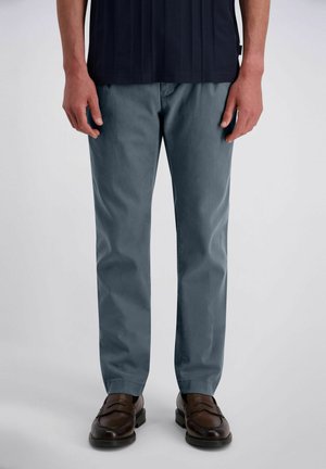 Personne portant un pantalon bleu droit et des mocassins en cuir marron, debout devant un fond clair uni.