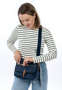 Junge Frau im gestreiften Hemd und Jeans lächelt, während sie eine marineblaue Crossbody-Tasche mit braunen Akzenten öffnet.