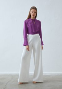 Lilla bluse med slipsnakke, parret med vide hvide bukser. Glat stof, klokkeærmer på blusen og minimalistisk design.