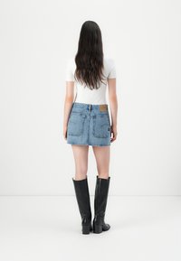 Ženska z dolgimi temnimi lasmi, oblečena v belo prilegajočo majico, modro denim mini krilo z zadnjimi žepi in črnimi škornji do kolen s peto, stoji ob belem ozadju.