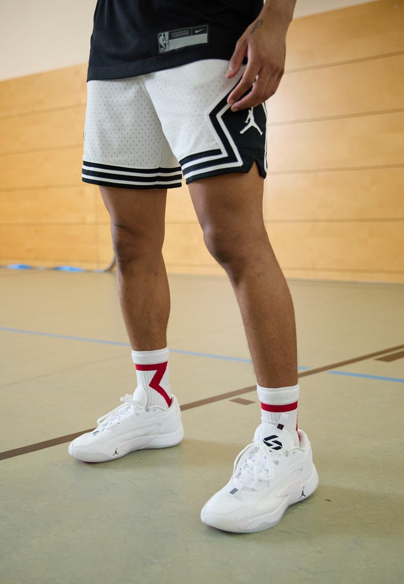 Weiße und schwarze Netz-Basketballshorts mit Streifen, kombiniert mit weißen Sneakern und Socken mit roten Akzenten. Indoor-Sportumgebung.