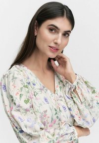 Kukk blouse, joka esittelee vaaleaa kermaista taustaa monivärisillä kukilla. Siinä on V-kaula-aukko ja puffihihat, joissa on sitomisdetaljit.