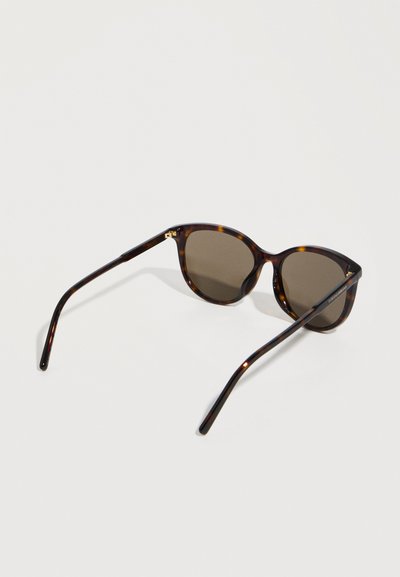 Γυαλιά ηλίου με καφέ σχέδιο πλαισίου τύπου χελώνας και σκοτεινούς φακούς, εμφανιζόμενα από πίσω πάνω σε ένα απλό ανοιχτόχρωμο υπόβαθρο.