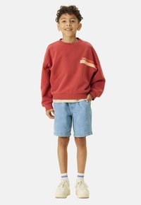 Sweatshirt rouge avec un design graphique, short en denim bleu clair avec un motif de visages souriants, et baskets beiges avec des accents blancs.