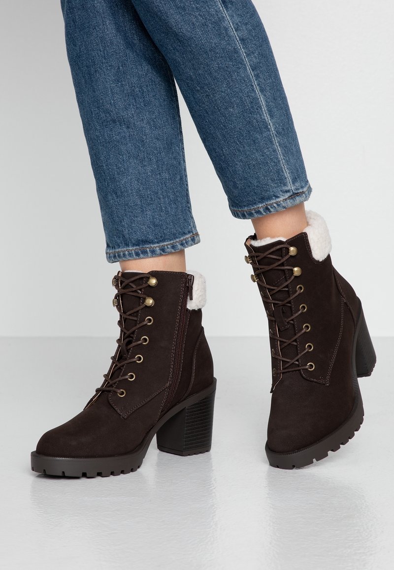 Bottes montantes en cuir marron foncé à lacets avec talons épais et doublure en polaire blanche, portées avec un jean bleu retroussé sur un sol réfléchissant.