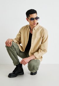 Beige gombos ing, fekete trikó, olíva zöld nadrág, fekete fűzős csizma, oversized napszemüveg, guggoló póz, minimalista háttér.