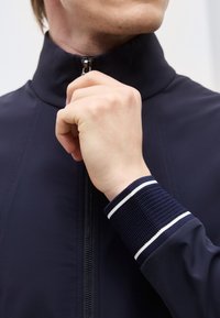 Το χέρι ενός άντρα κλείνει το φερμουάρ ενός ναυτικού μπλε μπουφάν με υφασμένο μανσέτα που έχει λευκές ρίγες.