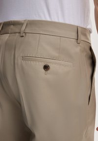 Beige broek met een gladde textuur, voorzien van een achterzak met een bruine knop. De stof heeft een subtiele glans. Opvallende slim fit.