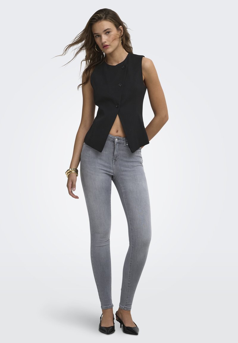 Jeune femme aux cheveux ondulés portant un haut noir sans manches à boutons, un jean skinny gris clair, des talons pointus noirs et des bracelets dorés.