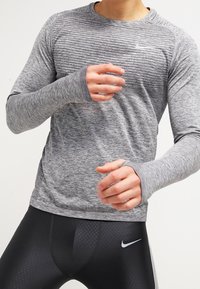 Grijze lange mouwen sportshirt met een gestructureerd ontwerp, met duimopeningen bij de manchetten. Gecombineerd met strakke zwarte leggings met een logo.