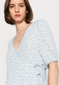 Vestido azul claro com decote em V e mangas curtas bufantes. Apresenta um padrão floral em verde e branco, com um detalhe de cordão de ajuste na lateral.