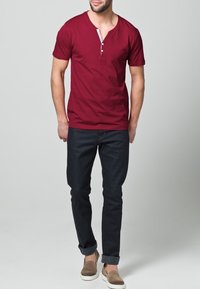 Bordové tričko s krátkým rukávem, výstřihem do V a třemi knoflíky, v kombinaci s tmavě modrými džínami a béžovými nazouvacími botami.