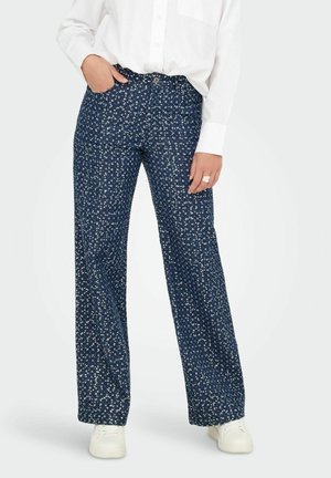 Persona che indossa pantaloni blu scuro con un motivo a piccoli punti bianchi, una camicia bianca con bottoni e sneakers bianche, in piedi con una mano in tasca.