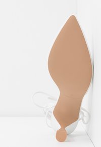 Chaussure à talon haut à bout pointu blanche inclinée contre un coin blanc, montrant une semelle lisse couleur bronzée et un talon élancé avec des lacets.