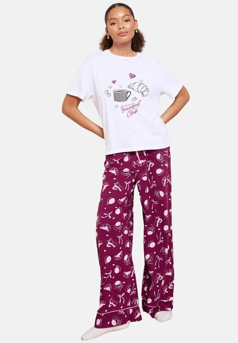 T-shirt bianco in cotone con grafica a tema colazione; pantaloni del pigiama viola con stampe di cibo e bevande in rosa. Vestibilità comoda, tessuto morbido, vita con cordoncino.