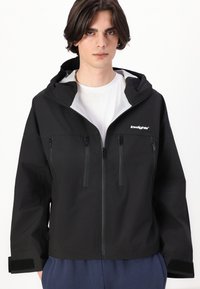 Veste noire imperméable avec capuche, fermeture éclair à l'avant, deux poches poitrine zippées et poignets avec bande Velcro. Texture lisse avec un détail de logo.