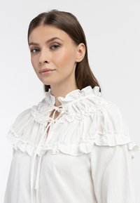 Blusa bianca con un alto colletto arricciato, chiusura con cordoncino e balze stratificate sulle maniche per un tocco di texture.